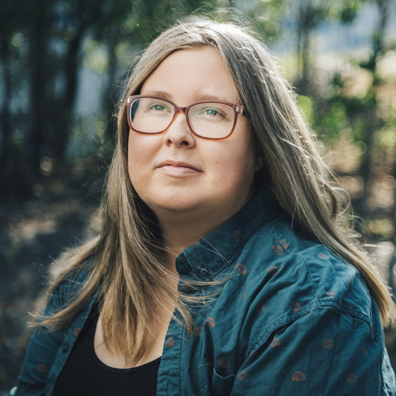 Portrait of Jacq Brasseur, a non-binary white person with long blonde hair and dark rimmed glasses. They are looking out of frame, to the left, with a small smile on their lips.