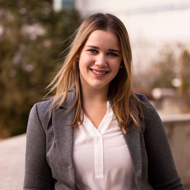 Portrait of Jacq Brasseur, a non-binary white person with long blonde hair and dark rimmed glasses. They are looking out of frame, to the left, with a small smile on their lips.