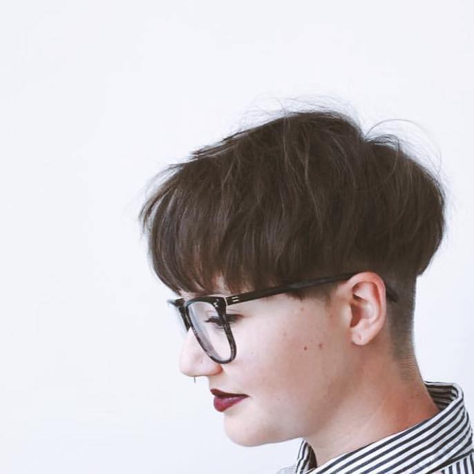 Portrait of Jacq Brasseur, a non-binary white person with long blonde hair and dark rimmed glasses. They are looking out of frame, to the left, with a small smile on their lips.