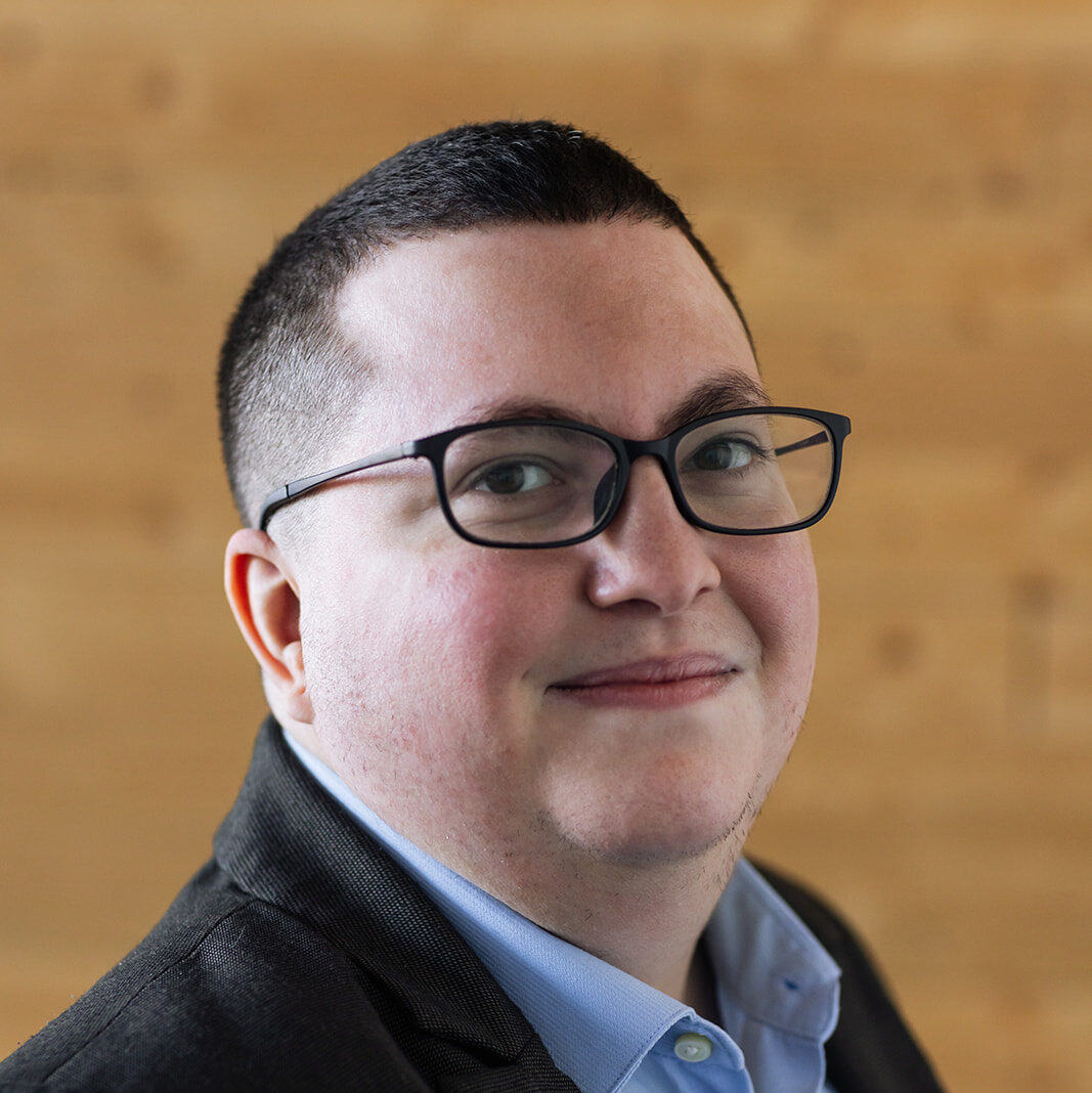 Portrait of Jacq Brasseur, a non-binary white person with long blonde hair and dark rimmed glasses. They are looking out of frame, to the left, with a small smile on their lips.